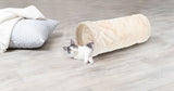 Weiß-graue Katze liegt in beige-rohrförmigem Kissen auf hellem Holzboden, neben einem weißen Kissen.
