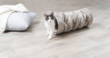 Eine schwarz-weiße Katze schaut aus einer flauschigen, grauen Röhre auf einem Holzfußboden, daneben ein graues Kissen.