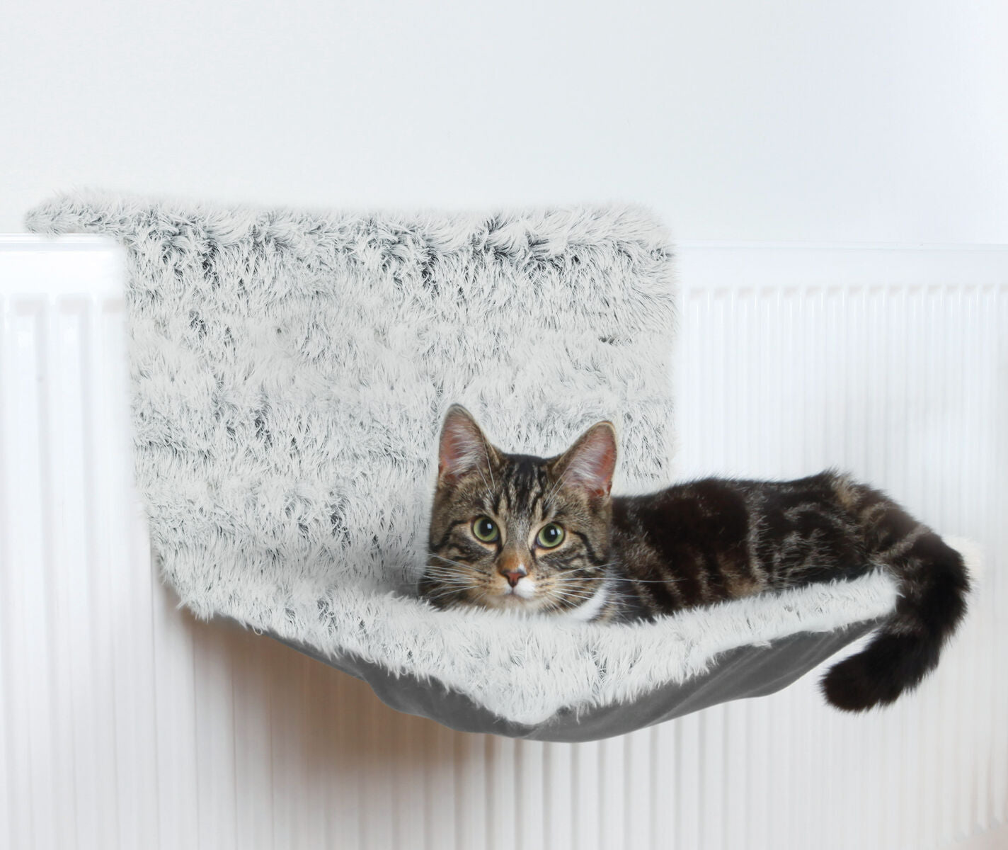 Eine getigerte Katze liegt auf einer grauen, pelzigen Hängematte, die an einem weißen Heizkörper angebracht ist.