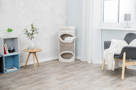 Helles Wohnzimmer mit Holzfußboden, ein grauer Sessel, ein runder Holztisch, ein Regal und ein Kratzbaum mit einer weißen Katze.