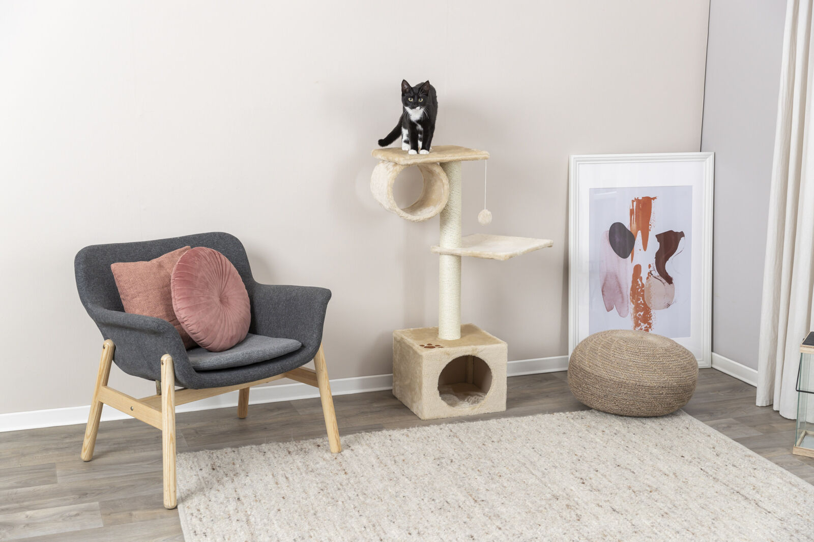 Katzenkratzbaum mit zwei Plattformen, auf dem eine schwarz-weiße Katze sitzt, daneben ein Sessel mit rosa Kissen.
