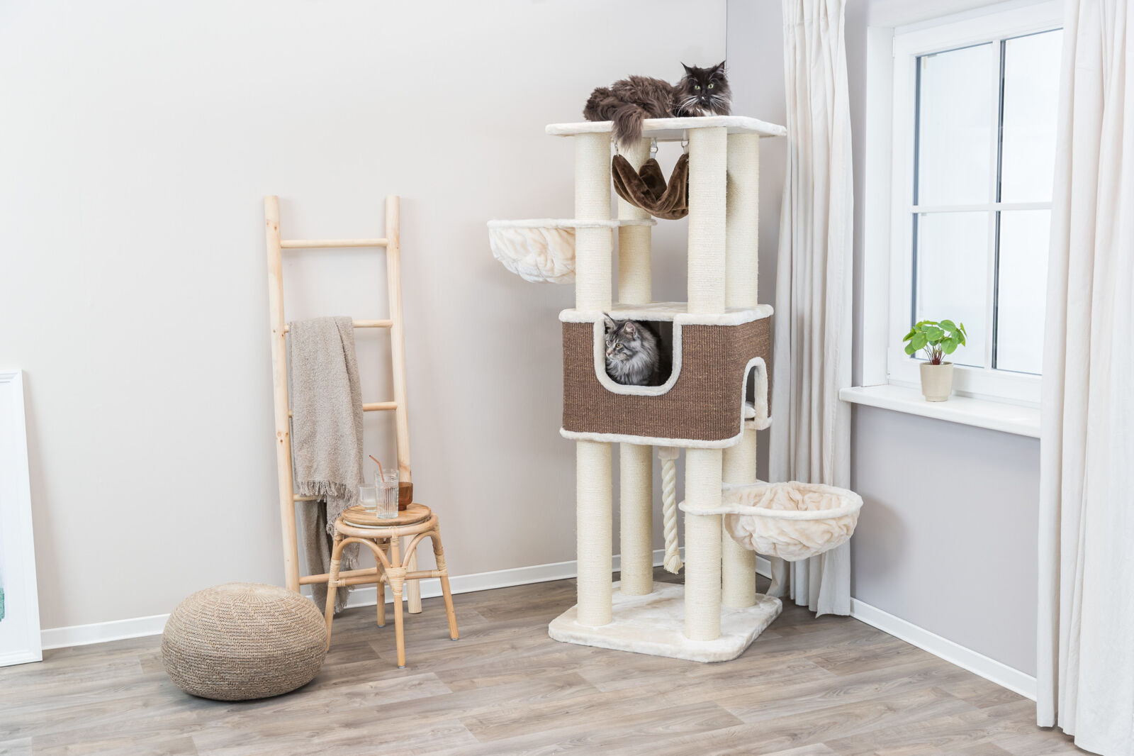 Katzenkratzbaum mit zwei Katzen, Stehleiter mit Decke, Hocker und Glas, Fensterbank mit Pflanze, helle Raumgestaltung.