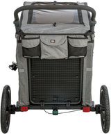 Ein grauer Fahrradanhänger mit zwei Rädern, oben mit Netzabdeckung und Griff, auf Asphalt stehend.