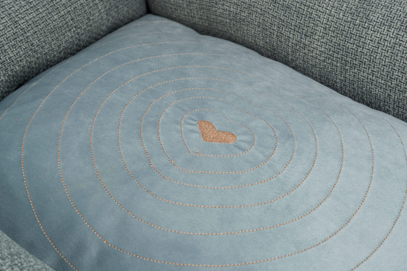 Graues Polsterkissen mit gesticktem Herzmuster in der Mitte und konzentrischen Kreisen auf einer Couch.