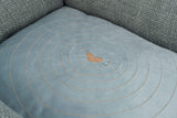 Graues Polsterkissen mit gesticktem Herzmuster in der Mitte und konzentrischen Kreisen auf einer Couch.