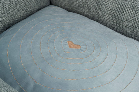 Graues Polsterkissen mit gesticktem Herzmuster in der Mitte und konzentrischen Kreisen auf einer Couch.