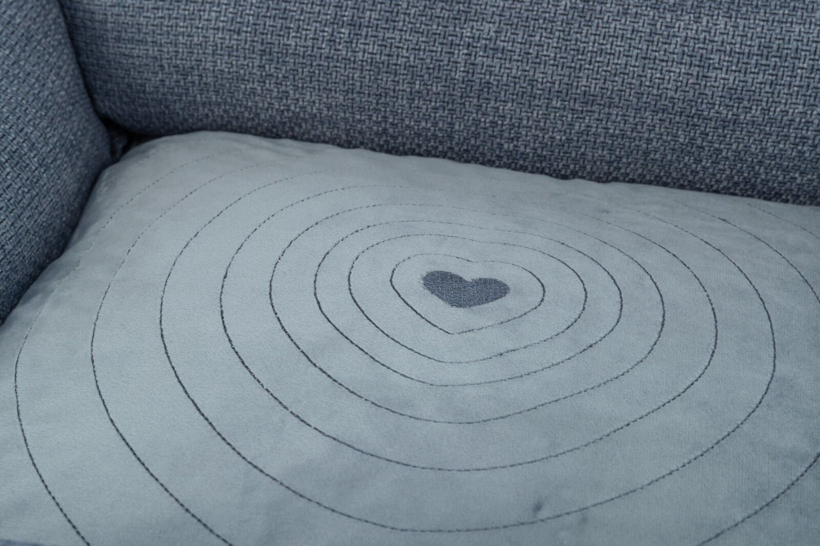 Graues Sofakissen mit gestickten konzentrischen Kreisen und einem Herz in der Mitte auf einem blauen Sofa.