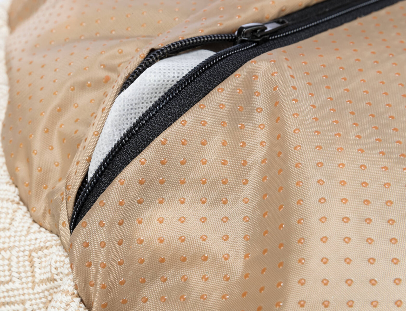 Nahaufnahme eines beigefarbenen Textils mit Punktmuster und einem schwarzen Reißverschluss, darunter ein weißes Mesh sichtbar.