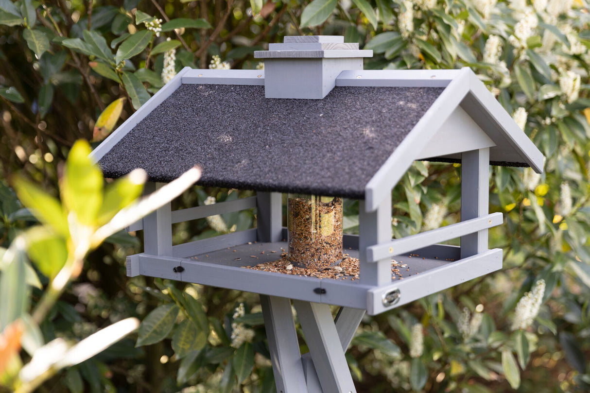 Graues Vogelhaus mit schwarzem Dach und Futterspender, umgeben von grünen Blättern und weißen Blüten.