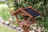 Holz-Vogelhaus mit schwarzem Dach steht im Garten, im Hintergrund sind grüne Pflanzen und Steine sichtbar.