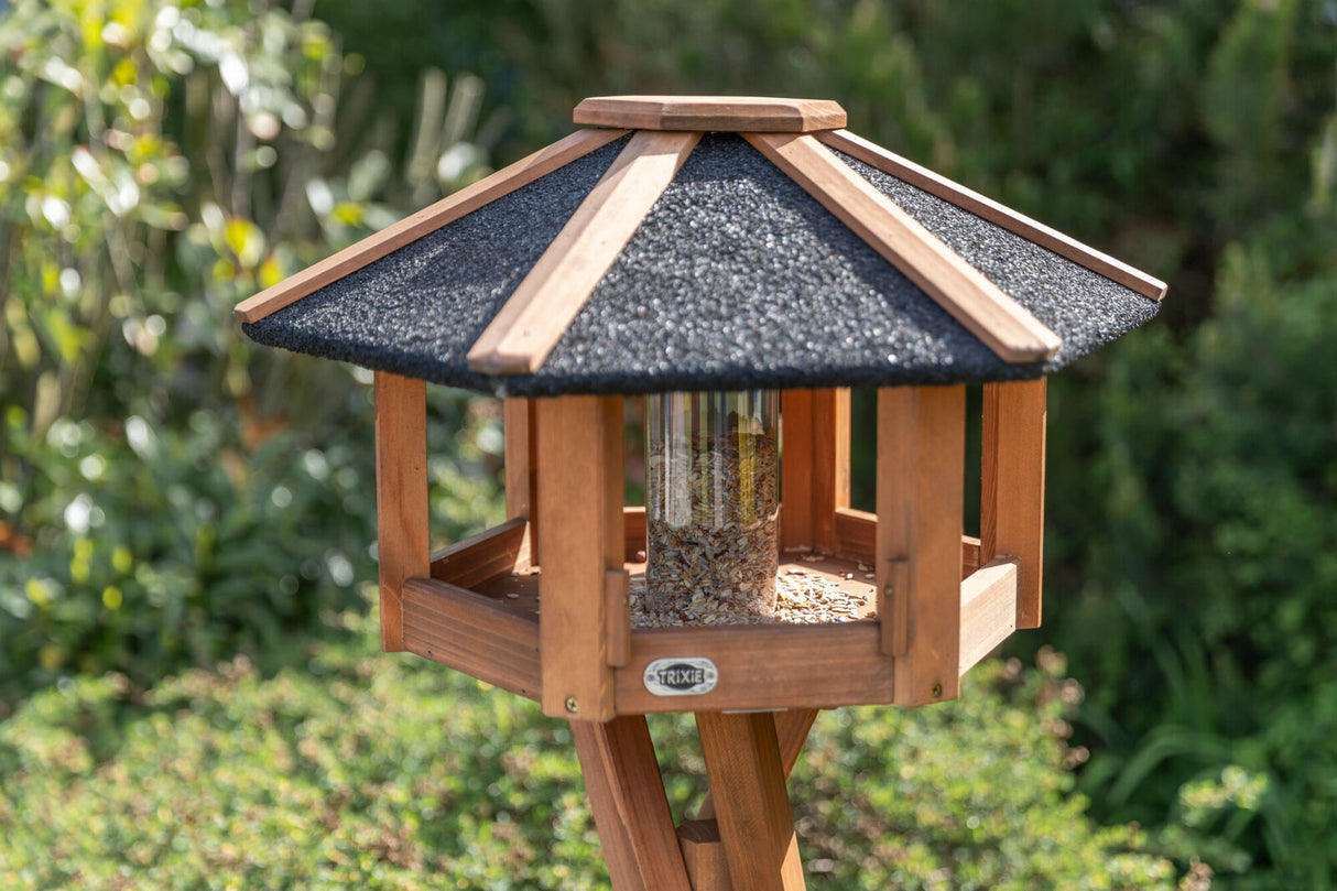 Sechseckiges Vogelhaus aus Holz mit schwarzem Dach, mittig gefüllter Glasbehälter mit Vogelfutter.