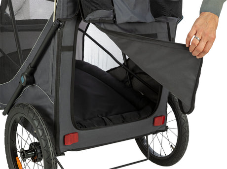 Ein grauer Fahrradanhänger mit Netzfenstern wird von einer Hand geöffnet, zeigt einen Innenraum mit Polsterung.