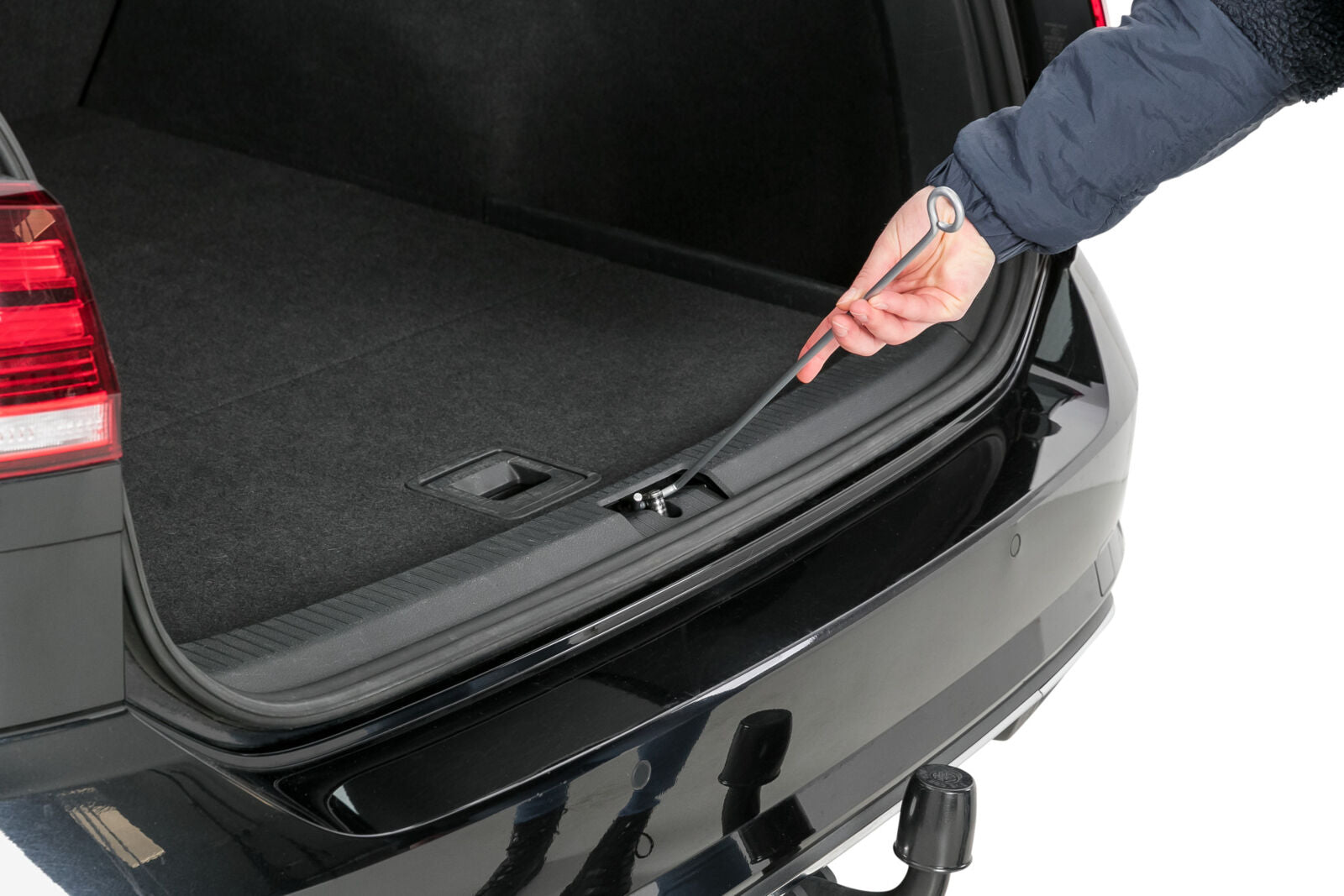 Eine Hand hebt mit einem Ringwerkzeug eine Abdeckung im Kofferraum eines schwarzen Autos an.