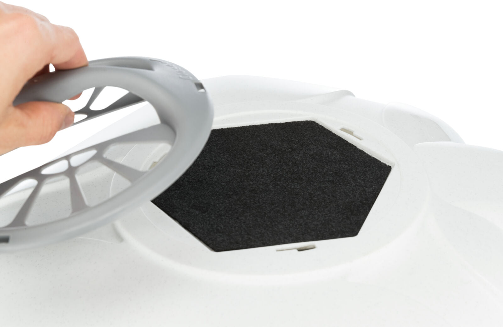 Hand hält graue Kunststoffabdeckung über einem schwarzen, sechseckigen Einsatz auf weißer Oberfläche.
