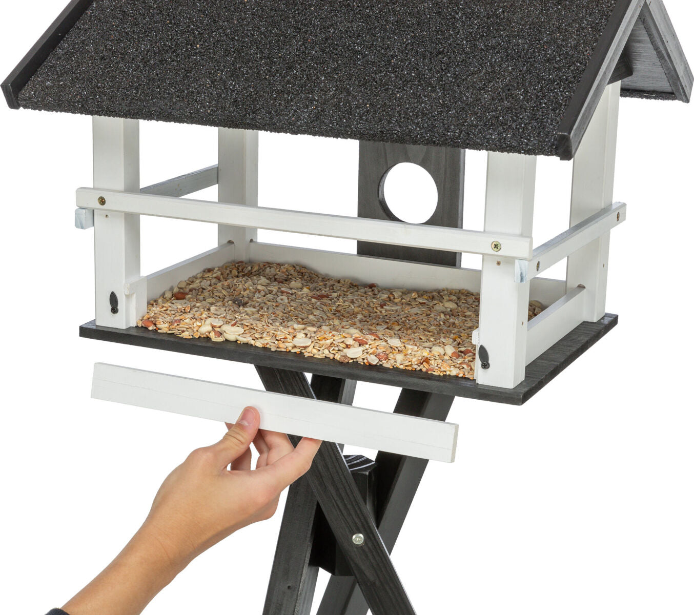 Vogelhaus mit schwarzem Dach und weißem Rahmen, gefüllt mit verschiedenen Samen, eine Hand hält ein Schiebebrett.