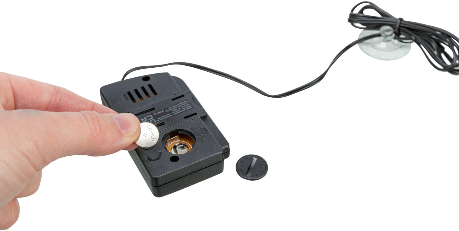 Eine Hand hält eine Knopfzelle, die vor einem schwarzen, verkabelten Gerät mit offenem Batteriefach positioniert ist, daneben liegt eine schwarze Abdeckung.