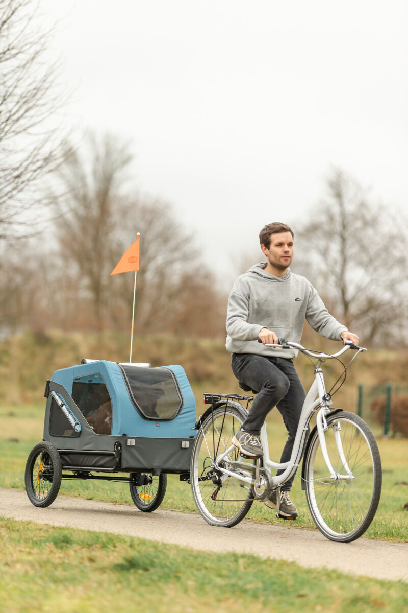 Mann auf Fahrrad zieht blauen Fahrradanhänger mit oranger Flagge auf ländlichem Weg.