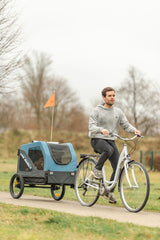 Mann auf Fahrrad zieht blauen Fahrradanhänger mit oranger Flagge auf ländlichem Weg.