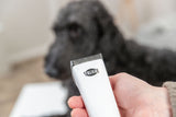 Eine Hand hält einen weißen Haarschneider mit der Aufschrift "Trixie" im Vordergrund, unscharfer schwarzer Hund im Hintergrund.