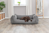 Hund liegt auf grauem Hundebett in einem Raum mit Holzboden, Pflanze und Fensterdeko.
