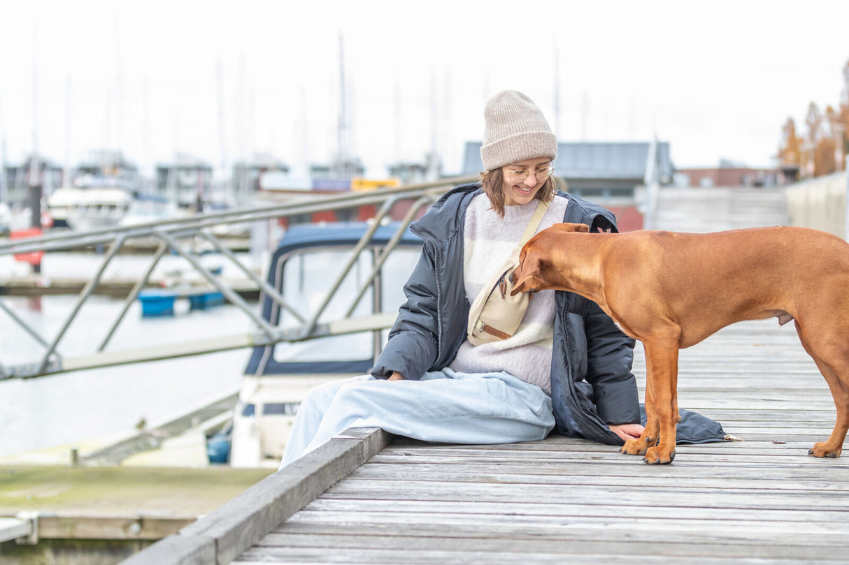 Eine Person sitzt lächelnd auf einem Holzsteg, während ein brauner Hund interessiert an einer Umhängetasche schnüffelt. Im Hintergrund sind Boote und Wasser zu sehen.