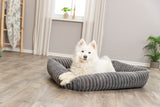 Ein weißer Hund liegt in einem grauen, gestreiften Hundebett auf einem Holzboden in einem hellen Raum.
