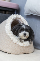 Ein kleiner Hund mit schwarz-weißem Fell liegt in einer weichen, beigen Höhle mit flauschigem Rand auf hellem Boden.