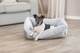 Hund liegt in einem grauen, flauschigen Hundebett auf Holzboden vor einem Sofa in neutralem Raum.