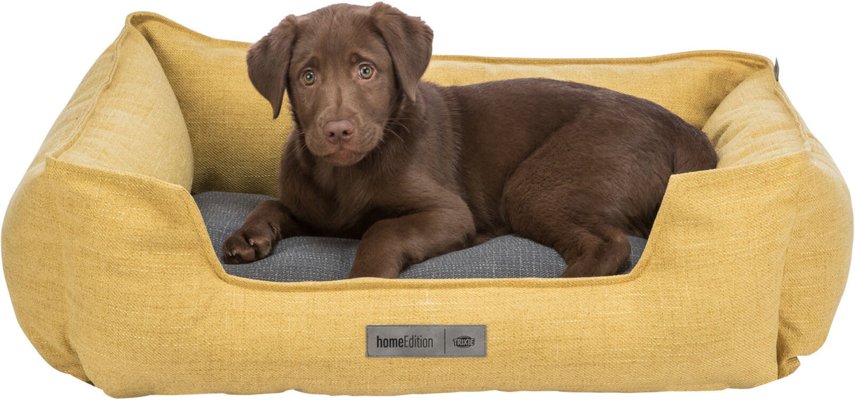 Ein brauner Welpe liegt in einem gelben Hundebett mit grauem Kissen, Aufschrift "homeEdition" und "TRIXIE".