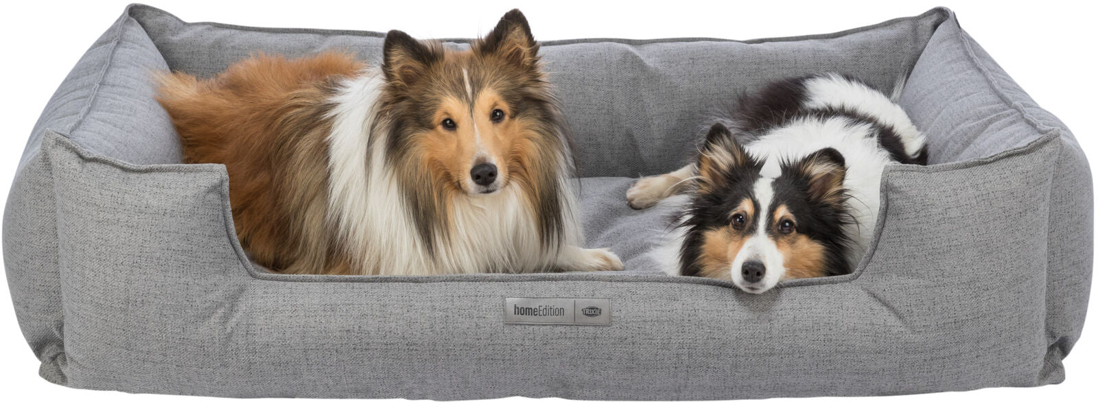Zwei Hunde liegen entspannt in einem grauen Hundebett mit einem Etikett, das "homeEdition TRIXIE" zeigt.