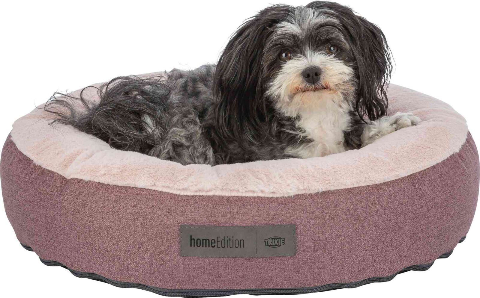 Kleiner langhaariger Hund liegt in einem runden, weichen, rosa Hundebett mit Label "homeEdition" und "TRIXIE".