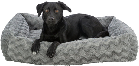 Ein schwarzer Hund liegt auf einem grauen, gemusterten Hundebett aus Plüsch und schaut nach vorne.