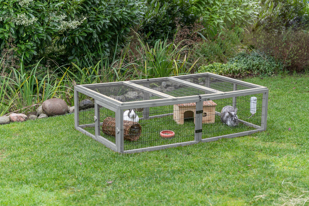 Rechteckiges Freigehege auf Rasen mit Kaninchen und Häuschen, umgeben von Pflanzen im Hintergrund.