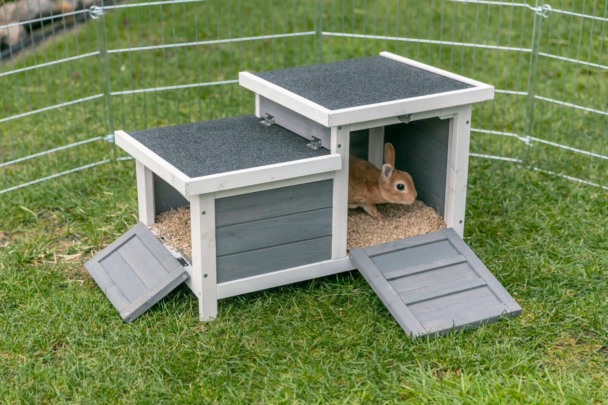 Grau-weiße Holzschutzhütte mit offenem Eingang, darin ein braunes Kaninchen auf hellbraunem Einstreu, auf grünem Gras.