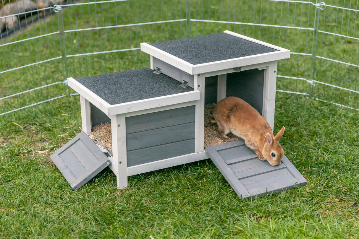 Holzhäuschen mit zwei Ebenen und schwarzem Dach steht auf Gras, ein braunes Kaninchen sitzt am Eingang.