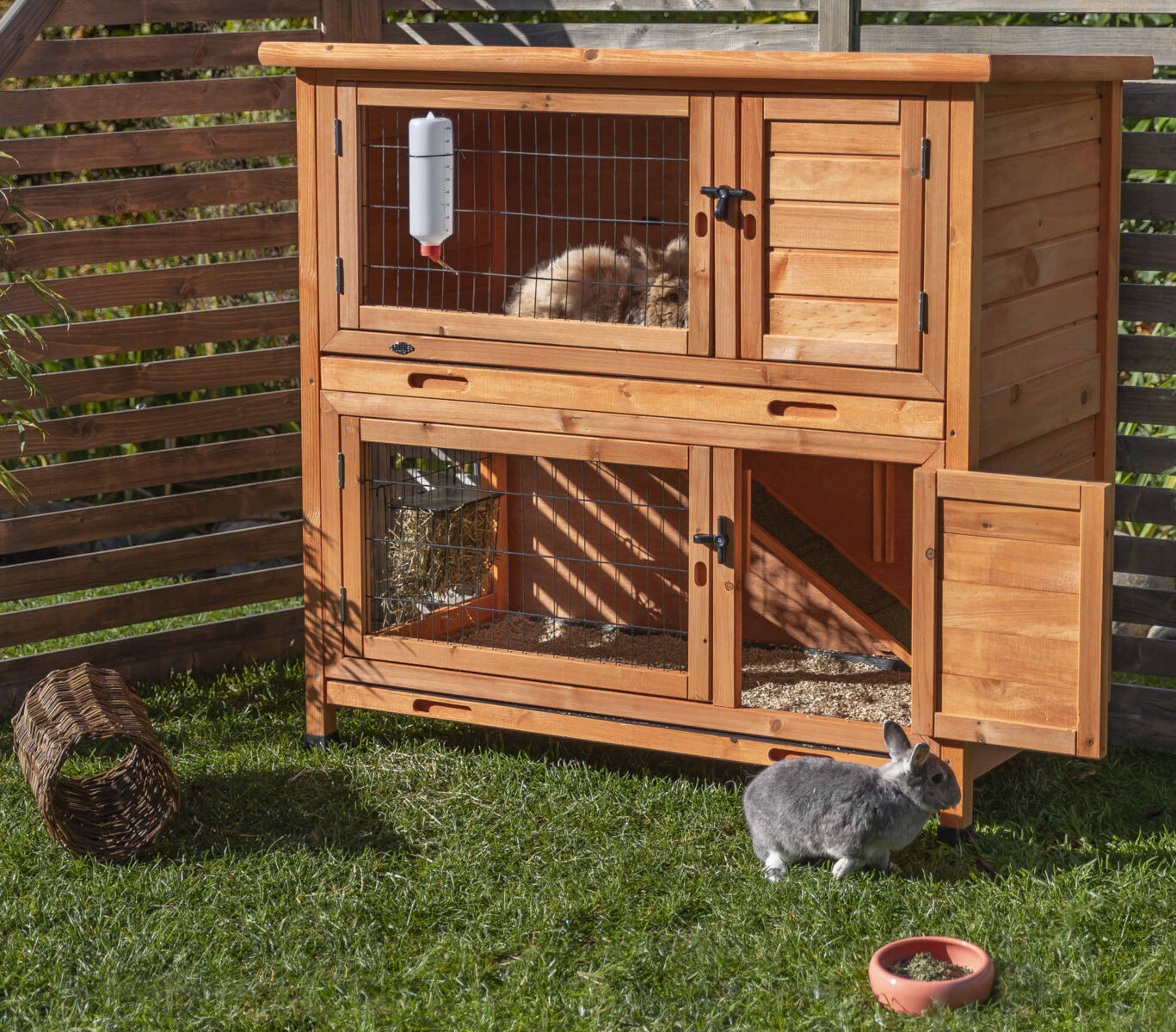 Holzkäfig mit zwei Etagen in einem Garten, oben ein Kaninchen und ein weiteres am Boden; Trinkflasche und Futternapf sichtbar.