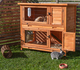 Holzkäfig mit zwei Etagen in einem Garten, oben ein Kaninchen und ein weiteres am Boden; Trinkflasche und Futternapf sichtbar.