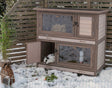 Holzkaninchenstall mit zwei Ebenen und offener Tür, ein Kaninchen innen, Schnee, Gräser und Bambuspflanze außen.