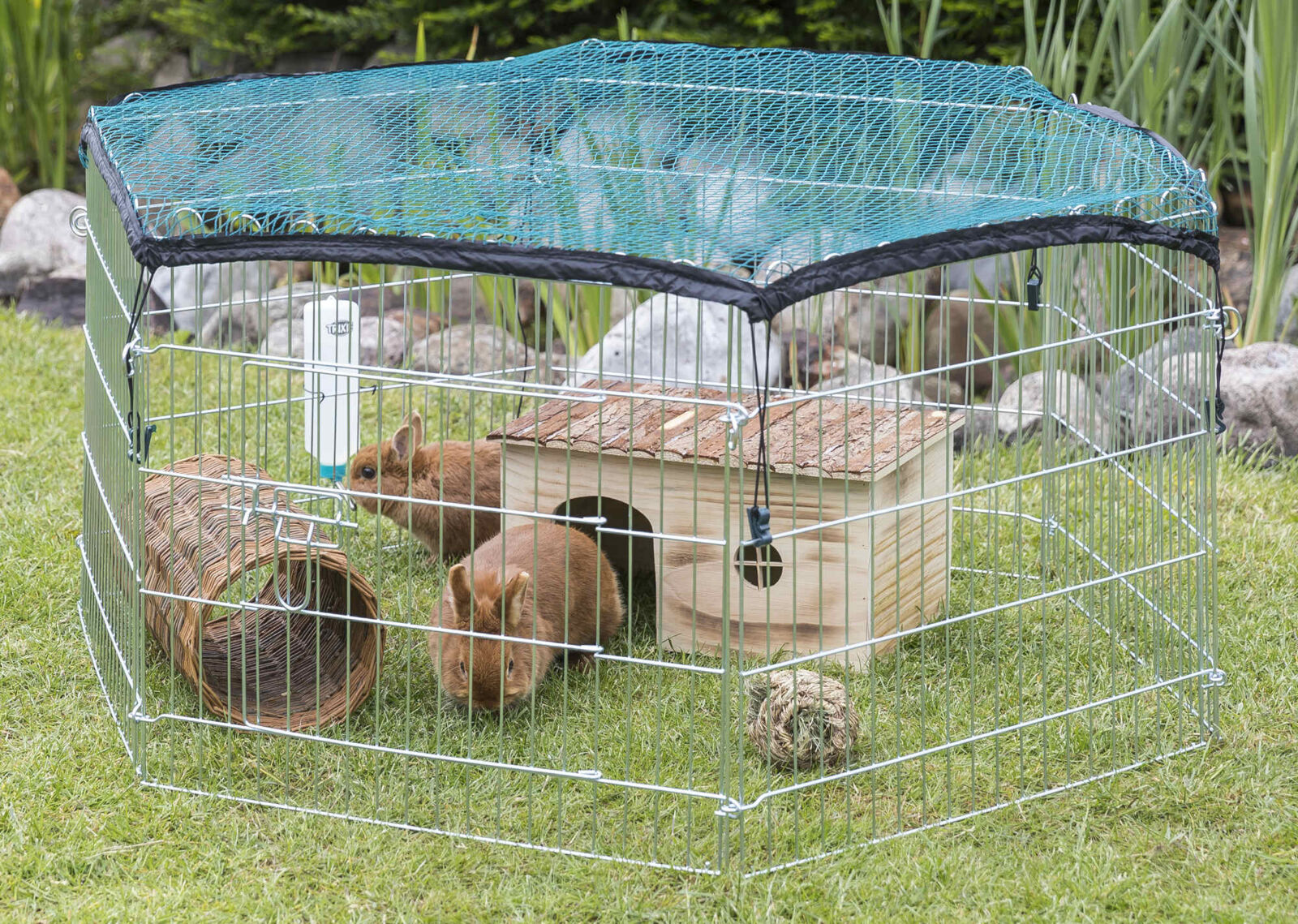 Drahtgehege mit grünem Netz für Kleintiere auf Gras, enthält zwei Kaninchen, Holzhaus und Weidentunnel.