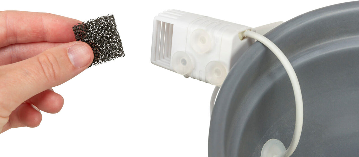 Hand hält schwarzes Netzstück vor einem weißen Plastikteil mit Saugnäpfen und einem Kabel, das an eine graue Oberfläche befestigt ist.