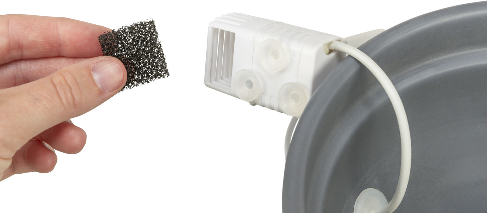 Hand hält schwarzes Netzstück vor einem weißen Plastikteil mit Saugnäpfen und einem Kabel, das an eine graue Oberfläche befestigt ist.