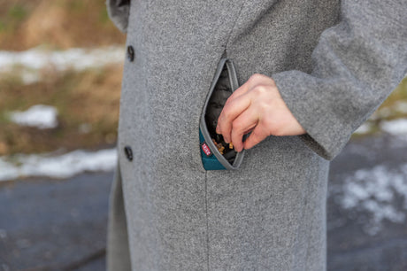 Eine Person in einem grauen Mantel steckt die Hand in eine Tasche, die etwas enthält, Hintergrund unscharf.