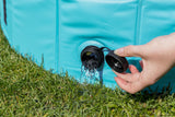 Eine Hand öffnet den Ablassverschluss eines blauen Wasserbeckens, Wasser fließt auf das grüne Gras.