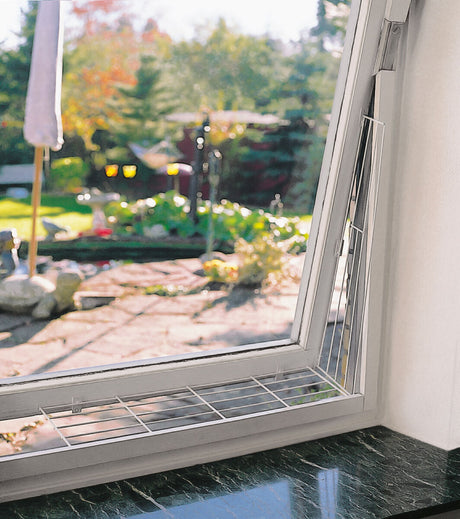 Offenes Fenster mit Gitter vor einem hellen, sonnigen Garten im Hintergrund; Gitter zur Sicherheit im unteren Fensterbereich befestigt.