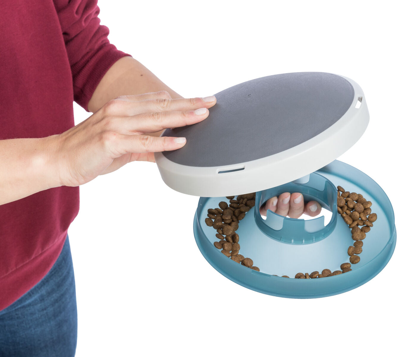 Eine Person in rotem Oberteil hält ein rundes, weißes und graues Futterspielzeug mit blauen Elementen und braunen Futterstücken.