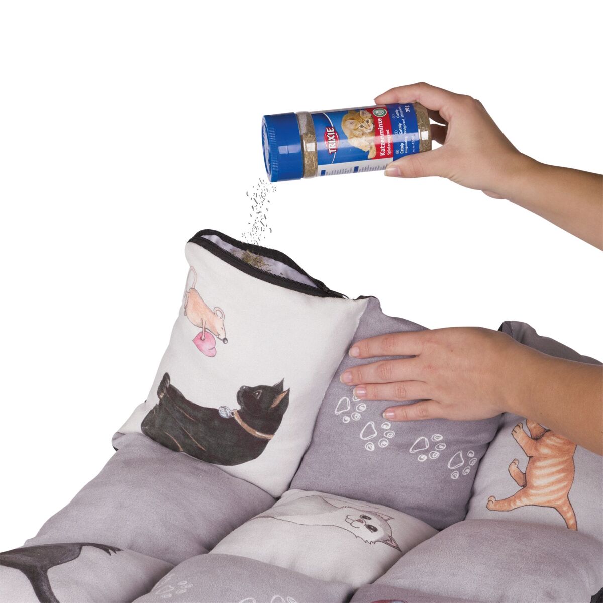 Person hält Behälter mit blauem Deckel und streut Pulver auf Kissen mit Katzenmotiven, graue und bunte Farben.