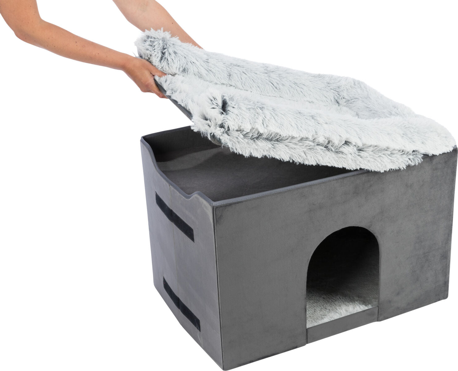 Eine Person hebt eine graue, flauschige Abdeckung von einer schwarzen rechteckigen Höhle mit einem Eingang an der Vorderseite.